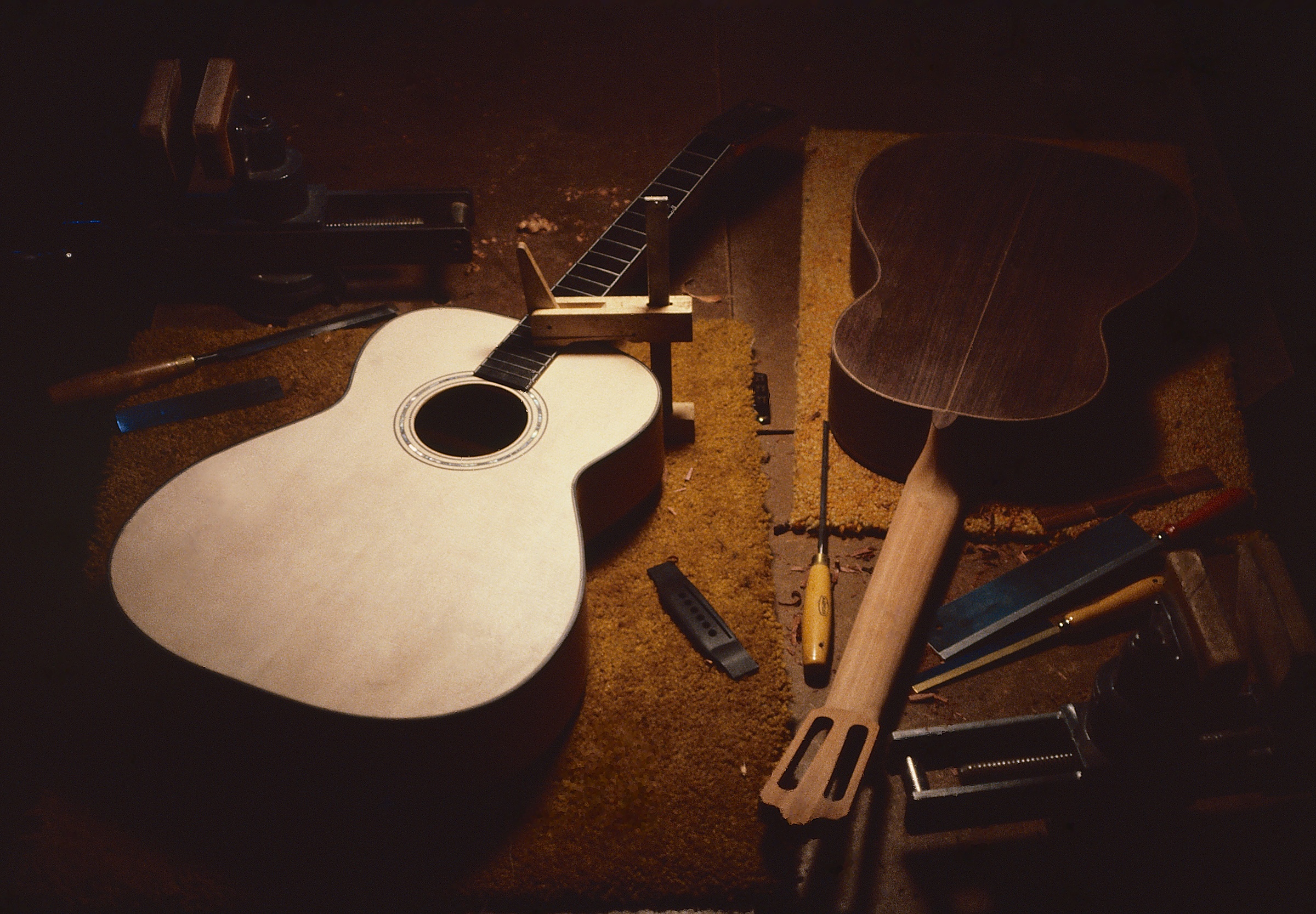 A spruce soundboard with its rosette inlaid and fingerboard fitted, a rosewood back and a carved mahogany neck laid out on a workbench among files, chisels and shavings